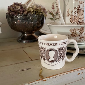 Könnte beinhalten: Eine weiße Keramiktasse mit einem braunen Design, das eine Silhouette von Königin Elisabeth II. und den Text "Queen's Silver Jubilee H.M. Queen Elizabeth II" zeigt.
