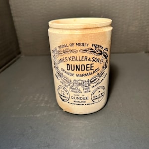 May include: An antique, off-white ceramic jar for orange marmalade. The jar features black text and illustrations, including "James Keiller & Son Dundee Orange Marmalade" and "Medal for Marmalade." The jar is cylindrical with a slightly flared rim.
