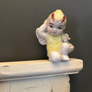 Könnte beinhalten: Eine kleine Porzellanfigur einer Fee mit gelben Flügeln und einem gelben und roten Kostüm. Die Fee sitzt auf einem weißen Regal.