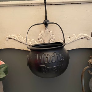 May include: Black cauldron with a handle hanging from a hook on a white shelf. The cauldron has the words "Witch's Brew" printed on the front.