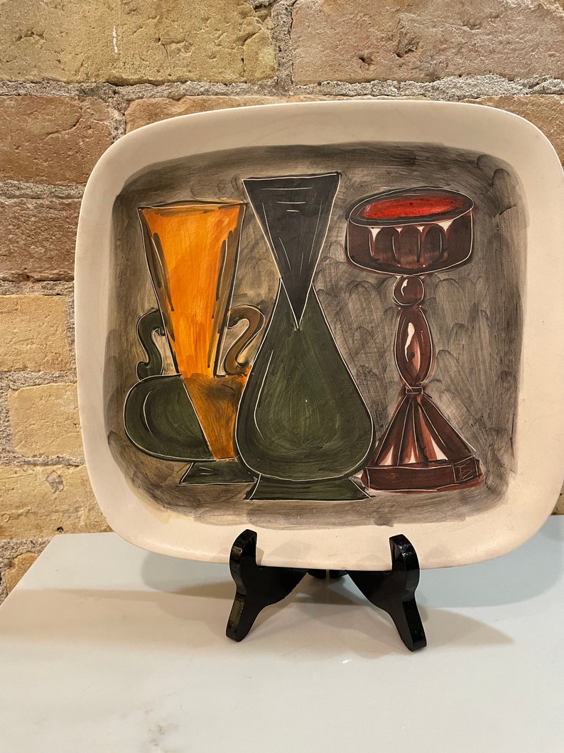May include: A ceramic serving tray with a hand-painted design of three stylized vases. The vases are painted in shades of green, orange, brown, and black against a gray background.