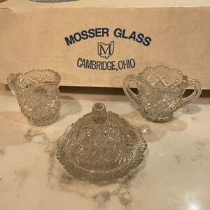 May include: A set of three clear glass pieces with a cut glass pattern. The pieces include a butter dish, a creamer, and a sugar bowl. The box they came in is visible in the background and says "Mosser Glass, Cambridge, Ohio".