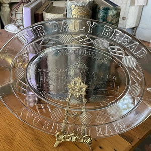 May include: A clear glass serving dish with a raised rim and handles. The dish is decorated with a repeating pattern of diamonds and circles. The centre of the dish features a raised image of a building with the text "Continental Hotel 1876". The rim of the dish is inscribed with the text "Give Us This Day Our Daily Bread".