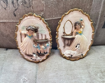 May include: Two oval-shaped wall plaques with a white background and gold trim. Each plaque features a 3D relief sculpture of a girl in a white dress with a dog. One plaque shows the girl holding a ball and the other shows the girl with three children.