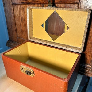 May include: An open, vintage wooden box with a reddish-brown exterior and a yellow interior. The lid is open, revealing a small mirror. The box has a brass clasp and a keyhole. The box is likely used for storing jewelry or small keepsakes.