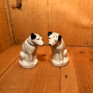 Puede incluir: Dos figuras de perro de cerámica. Uno tiene un collar negro, el otro manchas negras en las orejas. Ambos son blancos con orejas y nariz negras. Están sobre una superficie de madera.