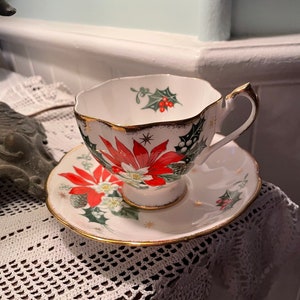 May include: A white teacup and saucer with a gold rim. The cup and saucer are decorated with a red poinsettia, green holly, and gold stars.