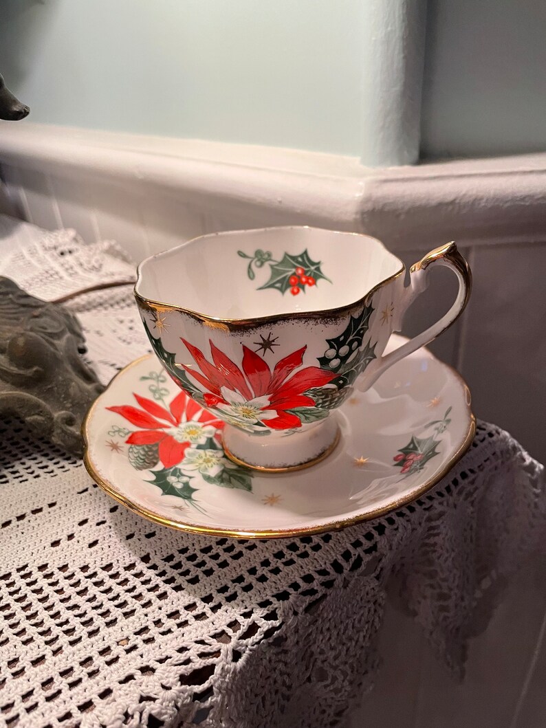 May include: A white teacup and saucer with a gold rim. The cup and saucer are decorated with a red poinsettia, green holly, and gold stars.