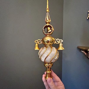 May include: A gold-toned metal tree topper featuring a central white swirled glass orb, a pointed finial, and three small hanging bells. The structure includes ornate scrollwork details and a hollow base designed for attachment to a tree branch.