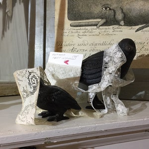 May include: Two black cast iron bird figurines with lace trim. The larger bird is standing on a shelf with a vintage print behind it. The smaller bird is sitting on the shelf in front of the larger bird. The print behind the birds has the text "Wahre und eigentliche An Ao1732. ohny" and "FICOTIN 199".