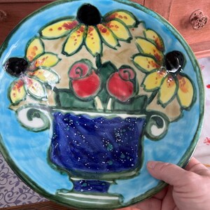 Op de afbeelding: Een blauw keramiek bord met een handgeschilderd ontwerp van gele bloemen in een blauwe en groene vaas. De bloemen hebben rode en zwarte centra.