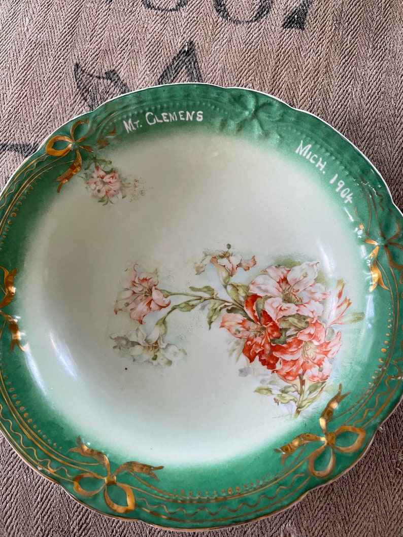 May include: A vintage green and white porcelain bowl with a floral design and gold trim. The bowl is inscribed with "Mt. Clemens Mich. 1904".