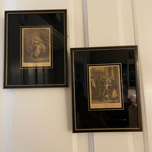 May include: Two framed black and gold prints with antique illustrations. The left print depicts a woman with a basket and two children. The right print depicts a group of people with a dog.