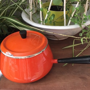 May include: Orange enamel fondue pot with a black handle and lid. The pot has a silver rim around the top.