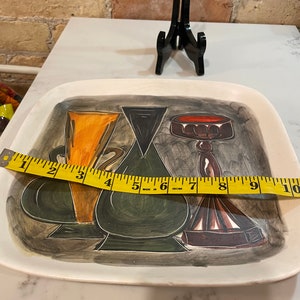 May include: A white ceramic serving tray with a painted design of three stylized vases. The vases are painted in shades of green, orange, brown, and red. The tray measures 10 inches in length.