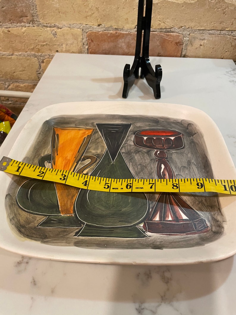 May include: A white ceramic serving tray with a painted design of three stylized vases. The vases are painted in shades of green, orange, brown, and red. The tray measures 10 inches in length.