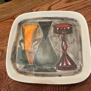 May include: A white ceramic serving platter with a hand-painted design of three stylized vases. The vases are painted in shades of black, green, orange, and brown. The platter has a square shape with rounded corners.