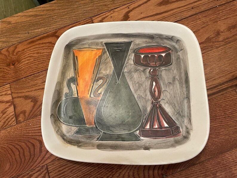 May include: A white ceramic serving platter with a hand-painted design of three stylized vases. The vases are painted in shades of black, green, orange, and brown. The platter has a square shape with rounded corners.
