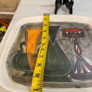 May include: A white ceramic serving tray with a black, green, and orange abstract design. The tray features three stylized vases with a red, brown, and black design. The tray measures 10 inches in length.