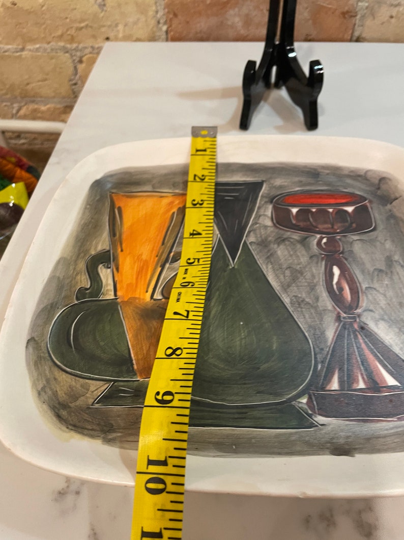 May include: A white ceramic serving tray with a black, green, and orange abstract design. The tray features three stylized vases with a red, brown, and black design. The tray measures 10 inches in length.