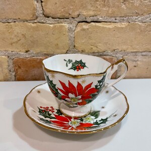 May include: A white teacup and saucer with a gold rim. The cup and saucer are decorated with a red poinsettia, green holly, and red berries. The design is on a white background with gold stars.