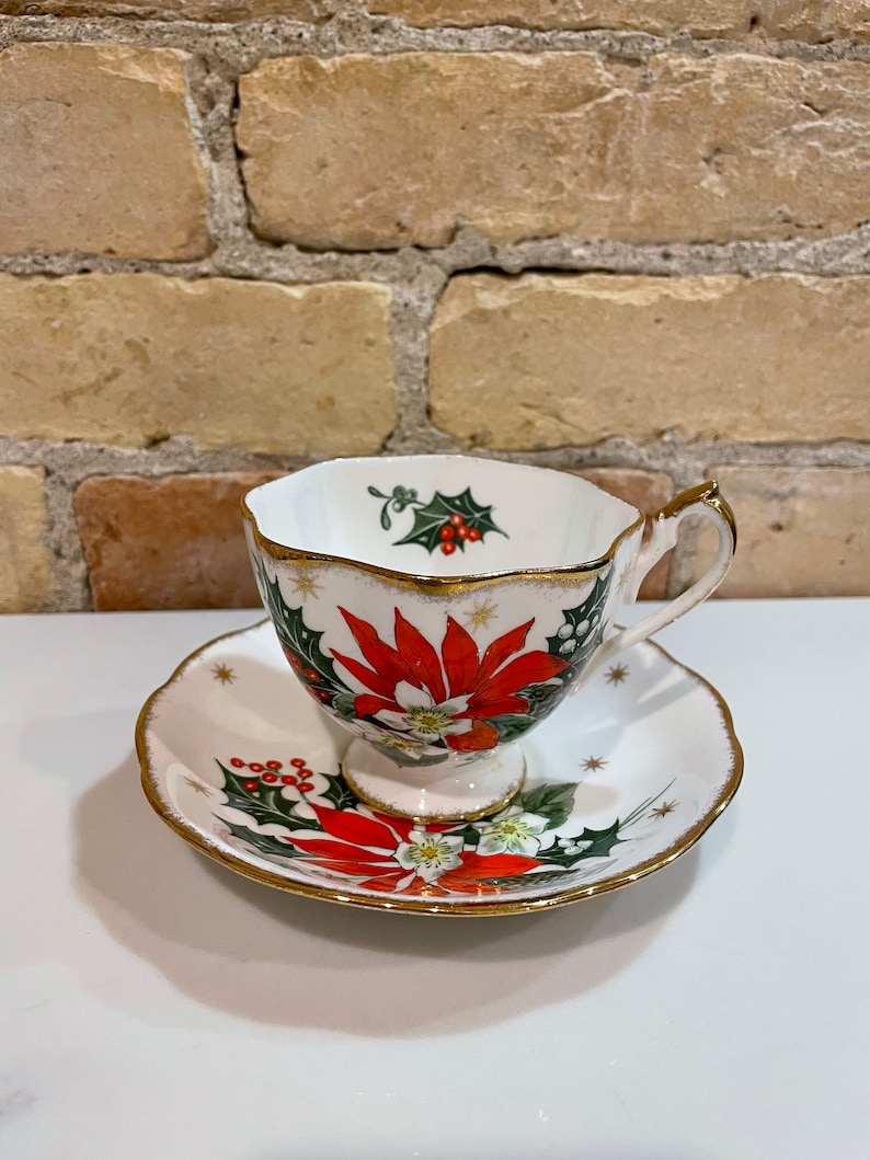 May include: A white teacup and saucer with a gold rim. The cup and saucer are decorated with a red poinsettia, green holly, and red berries. The design is on a white background with gold stars.