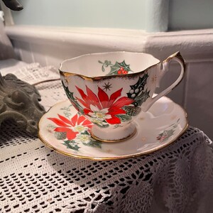 May include: A white teacup and saucer with a gold rim. The cup and saucer are decorated with a red poinsettia, green holly, and gold stars.