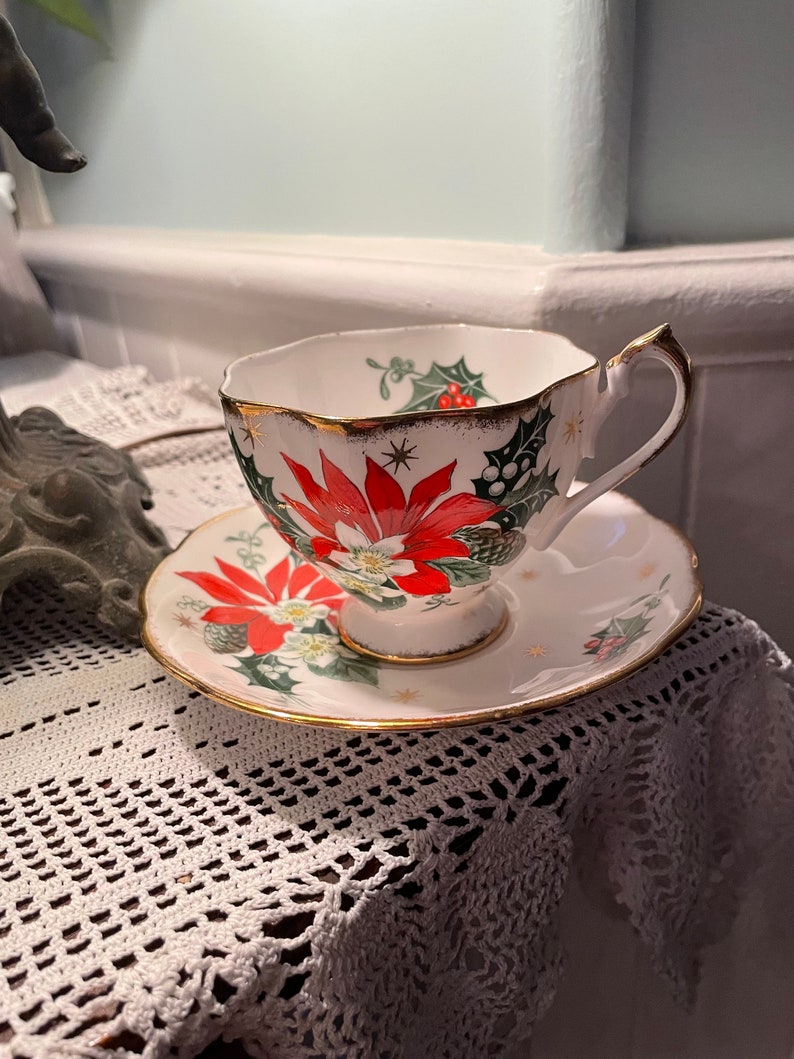 May include: A white teacup and saucer with a gold rim. The cup and saucer are decorated with a red poinsettia, green holly, and gold stars.