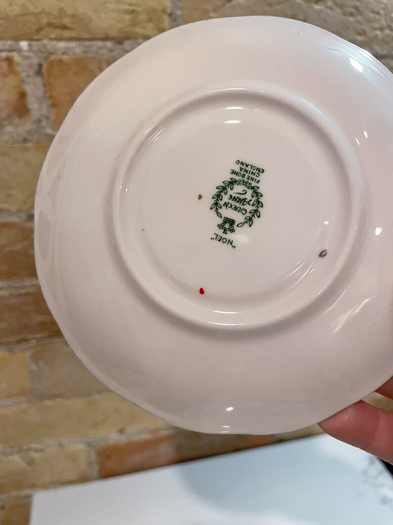 May include: White ceramic saucer with a green and gold design that reads "Queen Anne Fine Bone China England" and "Noel..."