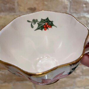 May include: A white teacup with a gold rim and a floral design. The cup features a green holly leaf with red berries.