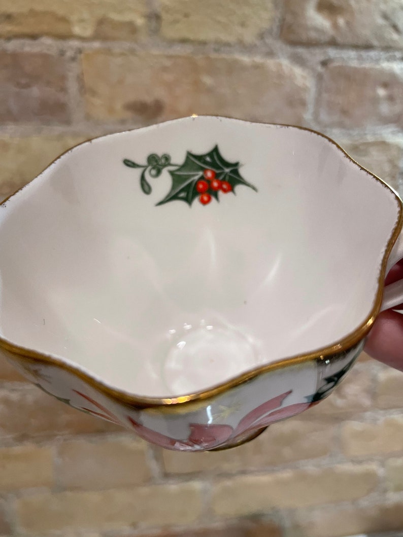 May include: A white teacup with a gold rim and a floral design. The cup features a green holly leaf with red berries.