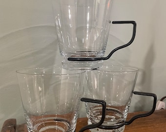 Op de afbeelding: Drie glazen bekers met zwarte metalen handvatten. De bekers zijn op elkaar gestapeld.