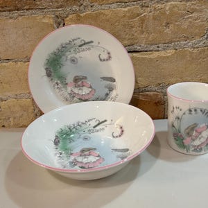 May include: A white ceramic dish set with a rabbit illustration in a pink dress, surrounded by flowers and greenery. The set includes a plate, a bowl, and a mug, all with pink rims.