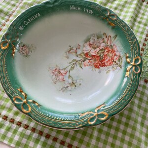 May include: A green and white porcelain bowl with a floral design and gold trim. The bowl has the words "Mt. Clemens Mich. 1904" written on the rim.