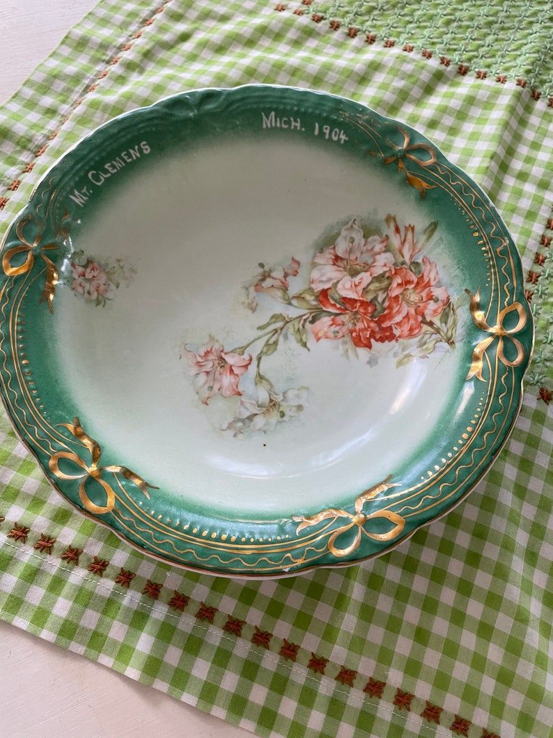 May include: A green and white porcelain bowl with a floral design and gold trim. The bowl has the words "Mt. Clemens Mich. 1904" written on the rim.