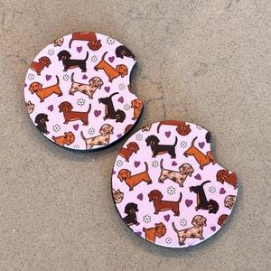 K&ouml;nnte beinhalten: Zwei runde Untersetzer in Rosa und Wei&szlig; mit einem Muster aus braunen, schwarzen und braunen Dackeln, Herzen und Blumen.