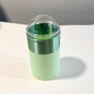 Może przedstawiać: Jasnozielony plastikowy kubek z przezroczystą, kopulastą pokrywką. Kubek ma metaliczny zielony pasek pośrodku. Kubek jest wypełniony ciemnozielonym płynem.