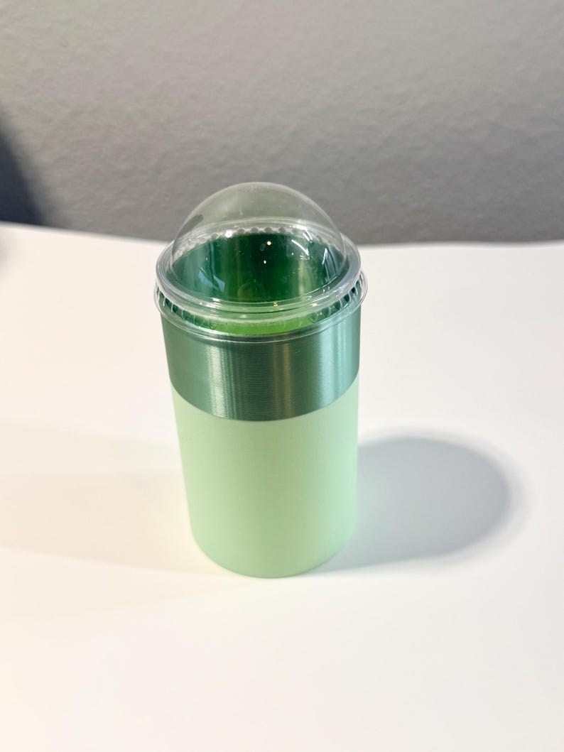 Może przedstawiać: Jasnozielony plastikowy kubek z przezroczystą, kopulastą pokrywką. Kubek ma metaliczny zielony pasek pośrodku. Kubek jest wypełniony ciemnozielonym płynem.