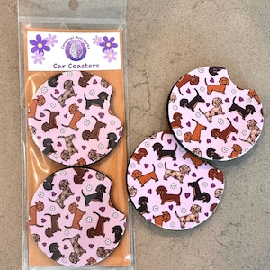K&ouml;nnte beinhalten: Set aus drei rosa Auto-Untersetzern mit einem sich wiederholenden Muster aus braunen und schwarzen Dackeln mit wei&szlig;en Blumen und lila Herzen. Die Untersetzer sind in einer durchsichtigen Plastikt&uuml;te mit einem Etikett verpackt, auf dem "Cerebral Artwork Car Coasters" steht.
