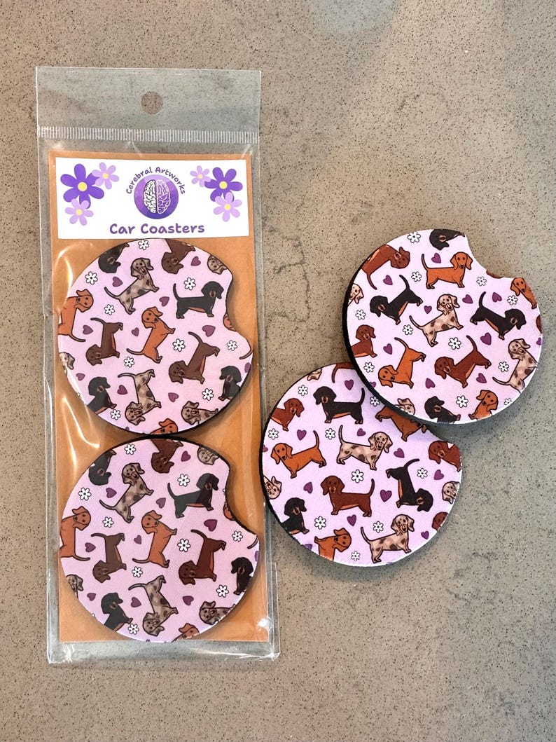 K&ouml;nnte beinhalten: Set aus drei rosa Auto-Untersetzern mit einem sich wiederholenden Muster aus braunen und schwarzen Dackeln mit wei&szlig;en Blumen und lila Herzen. Die Untersetzer sind in einer durchsichtigen Plastikt&uuml;te mit einem Etikett verpackt, auf dem "Cerebral Artwork Car Coasters" steht.