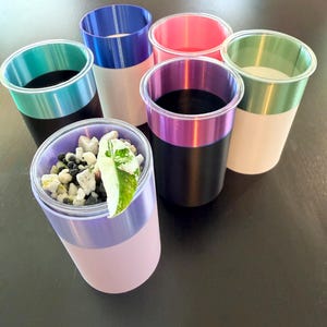 Może przedstawiać: Kolekcja dwukolorowych plastikowych kubk&oacute;w w r&oacute;żnych kolorach. Obręcze są w kolorze turkusowym, niebieskim, r&oacute;żowym, fioletowym i zielonym. Jeden kubek zawiera małe kamienie i zielono-biały liść. Kubki są ułożone na ciemnej powierzchni.