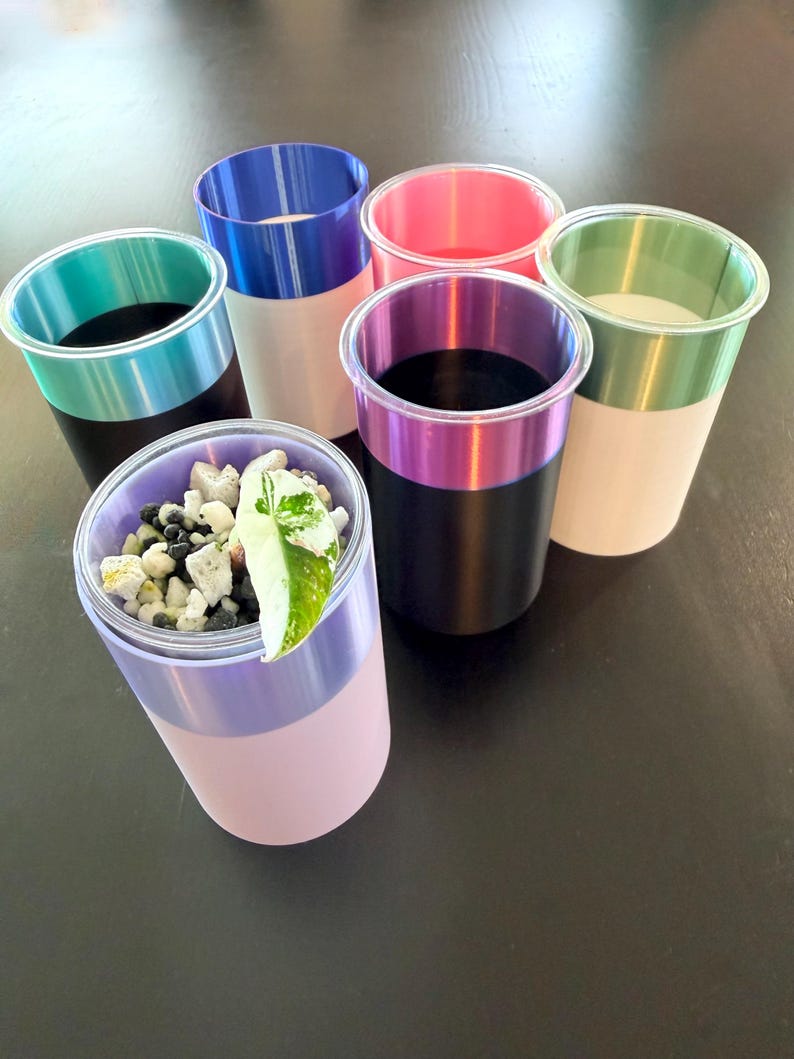 Może przedstawiać: Kolekcja dwukolorowych plastikowych kubk&oacute;w w r&oacute;żnych kolorach. Obręcze są w kolorze turkusowym, niebieskim, r&oacute;żowym, fioletowym i zielonym. Jeden kubek zawiera małe kamienie i zielono-biały liść. Kubki są ułożone na ciemnej powierzchni.