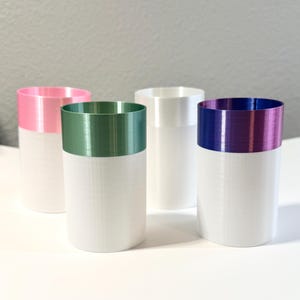 Może przedstawiać: Cztery cylindryczne wazony z białą podstawą i kolorowymi obrzeżami. Obrzeża są r&oacute;żowe, zielone, białe i fioletowe. Wazony ustawione są na białej powierzchni na szarym tle. Wazony są prawdopodobnie przeznaczone do dekoracji wnętrz.