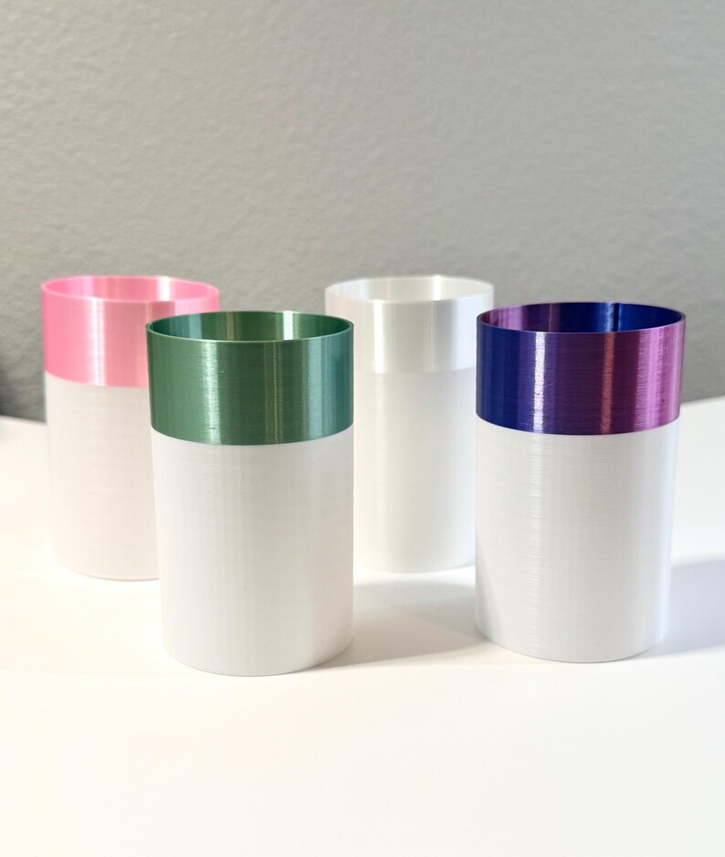 Może przedstawiać: Cztery cylindryczne wazony z białą podstawą i kolorowymi obrzeżami. Obrzeża są r&oacute;żowe, zielone, białe i fioletowe. Wazony ustawione są na białej powierzchni na szarym tle. Wazony są prawdopodobnie przeznaczone do dekoracji wnętrz.