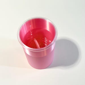 Może przedstawiać: R&oacute;żowa świeca w przezroczystym plastikowym kubku. Wosk świecy jest w żywym r&oacute;żowym kolorze, a biały knot jest widoczny na środku. Kubek ma matową r&oacute;żową podstawę i przezroczystą krawędź.