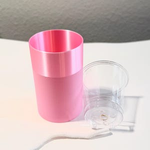 Może przedstawiać: R&oacute;żowy cylindryczny pojemnik z jaśniejszym r&oacute;żowym paskiem na g&oacute;rze, obok przezroczystego plastikowego kubka i białego sznurka. Obiekty znajdują się na białej powierzchni.