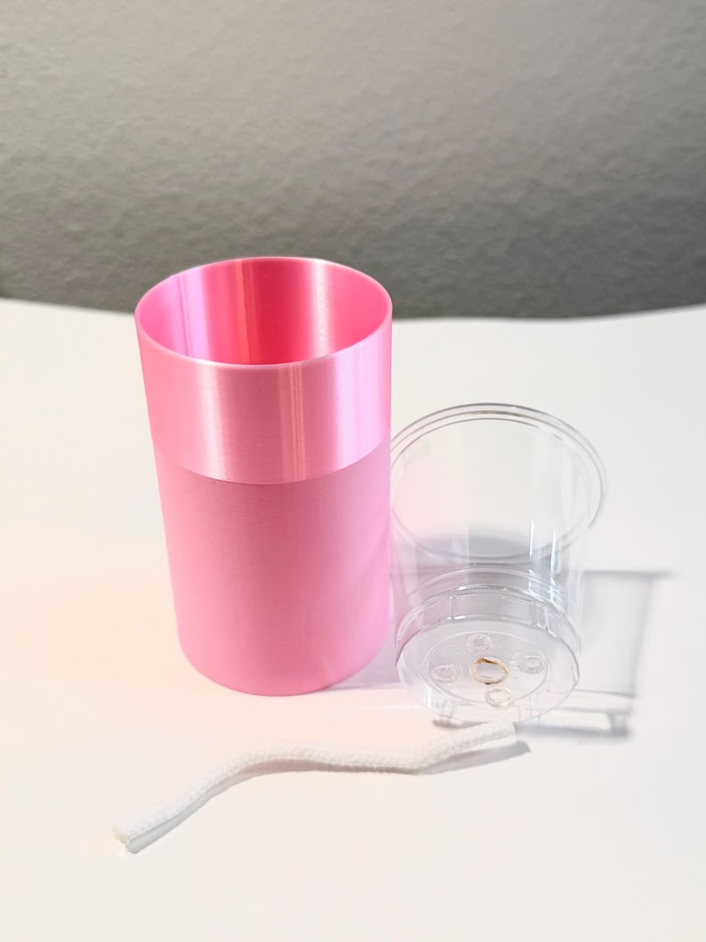 Może przedstawiać: R&oacute;żowy cylindryczny pojemnik z jaśniejszym r&oacute;żowym paskiem na g&oacute;rze, obok przezroczystego plastikowego kubka i białego sznurka. Obiekty znajdują się na białej powierzchni.