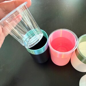 Może przedstawiać: Ręka trzyma przezroczysty plastikowy kubek nad czarnym kubkiem z turkusowo-fioletową obw&oacute;dką. Widoczne są inne kubki, w tym r&oacute;żowe i białe kubki z kolorowymi obw&oacute;dkami. Kubki znajdują się na czarnej powierzchni.