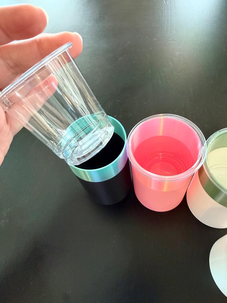 Może przedstawiać: Ręka trzyma przezroczysty plastikowy kubek nad czarnym kubkiem z turkusowo-fioletową obw&oacute;dką. Widoczne są inne kubki, w tym r&oacute;żowe i białe kubki z kolorowymi obw&oacute;dkami. Kubki znajdują się na czarnej powierzchni.