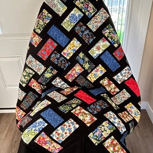 May include: A black and white quilt with a patchwork design featuring floral patterns in shades of red, blue, and green. The quilt is folded and draped over a chair.